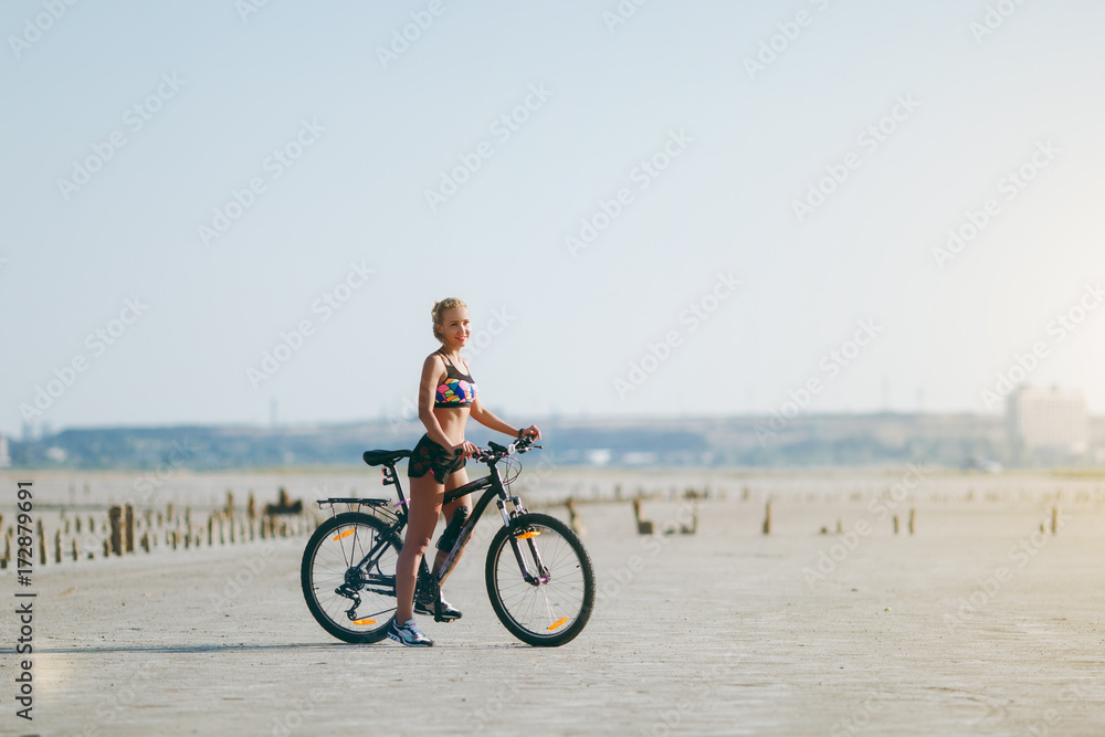 A strong blonde woman in a multicolored suit sits on a bicycle in a desert area and looks at the sun. Fitness concept. Blue sky background