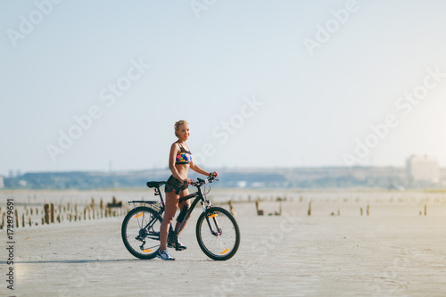 A strong blonde woman in a multicolored suit sits on a bicycle in a desert area and looks at the sun. Fitness concept. Blue sky background