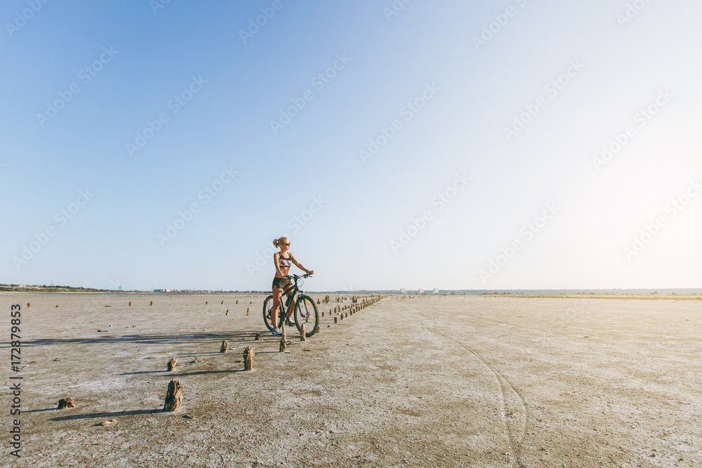 Fototapeta premium A strong blonde woman in a multicolored suit and sunglasses sits on a bicycle in a desert area and looks at the sun. Fitness concept. Blue sky background