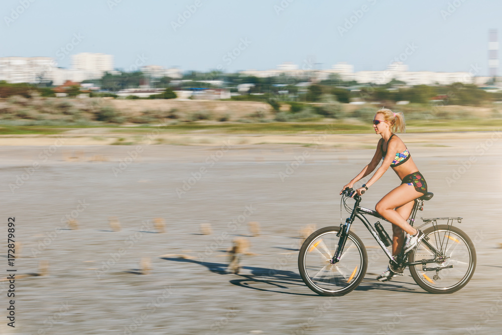 Fototapeta premium A sporty blonde woman in a colorful suit rides a bike at fast speed in a desert area on a sunny summer day. Fitness concept. Blur background