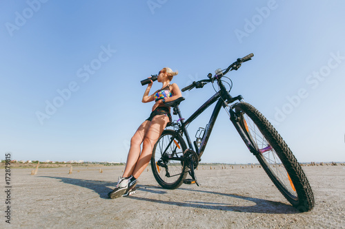 A strong blonde woman in a multicolored suit and sunglasses stands near a bicycle, drinks water from a bottle in a desert area. Fitness concept. Blue sky background