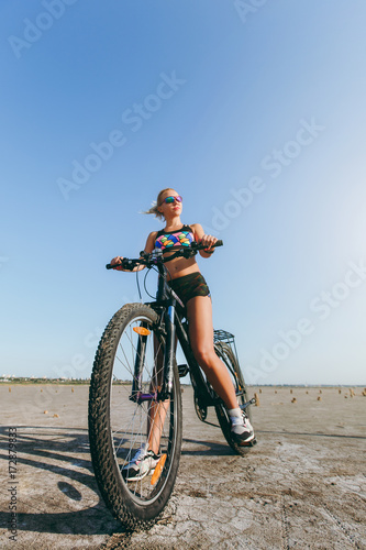 A strong blonde woman in a multicolored suit and sunglasses sits on a bicycle in a desert area and looks at the sun. Fitness concept. Blue sky background