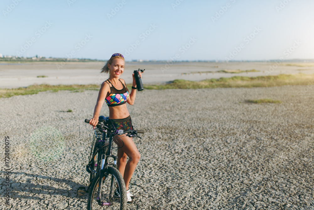 Fototapeta premium A strong blonde woman in a colorful suit and sunglasses stands near a bicycle with black bottle of water in a desert area. Fitness concept.
