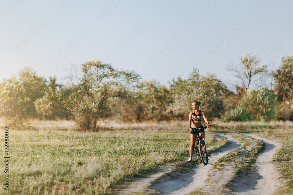 A strong blonde woman in a colorful suit sits on a bicycle in a desert area with trees and green grass and looks at the sun. Fitness concept. A strong blonde woman in a colorful suit sits on a bicycle in a desert area with trees and green grass and looks at the sun. Fitness concept.