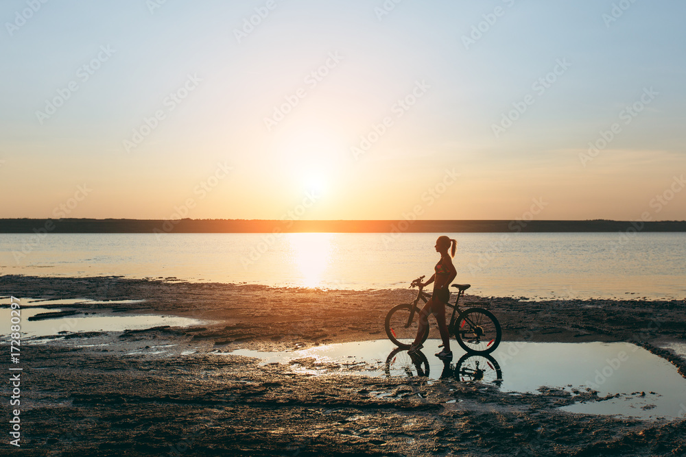 Fototapeta premium A strong blonde woman in a colorful suit stands near the bicycle in the water at sunset on a warm summer day. Fitness concept. Sky background