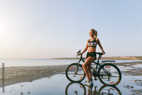 A strong blonde woman in a colorful suit stands near a bicycle in a desert area near the water. Fitness concept. Blue sky background