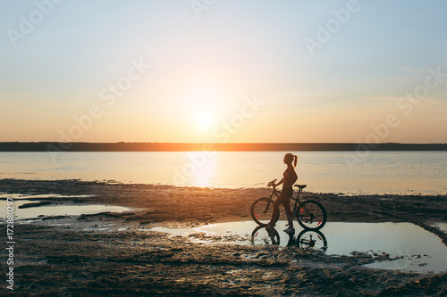 A strong blonde woman in a colorful suit stands near the bicycle in the water at sunset on a warm summer day. Fitness concept. Sky background © ViDi Studio