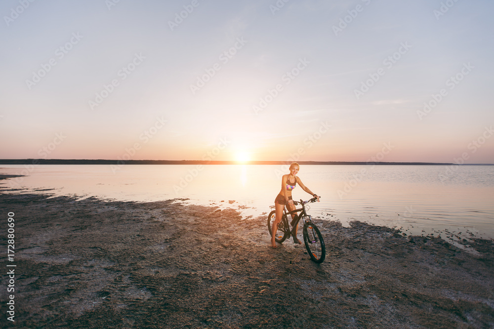 Fototapeta premium A sporty blonde woman in a colorful suit rides a bike in a desert area near the water on a sunny summer day. Fitness concept. Blue sky background
