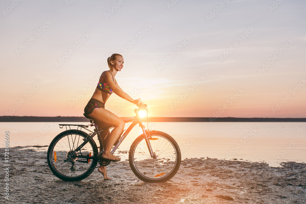 Fototapeta premium A sporty blonde woman in a colorful suit rides a bike in a desert area near the water on a sunny summer day. Fitness concept. Blue sky background