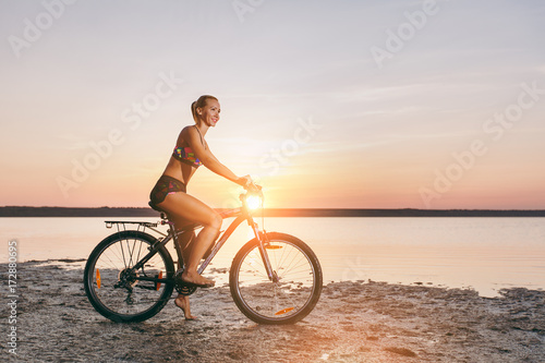 A sporty blonde woman in a colorful suit rides a bike in a desert area near the water on a sunny summer day. Fitness concept. Blue sky background