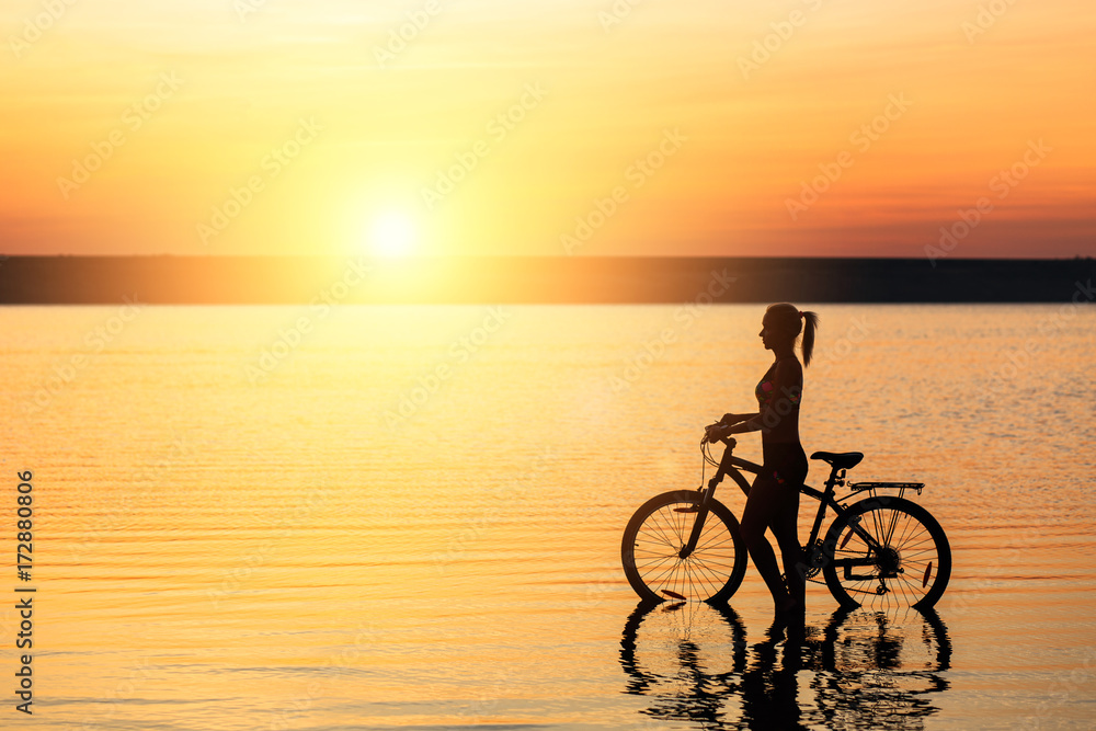 Obraz premium Silhouette of a sporty girl in a suit that sits on a bicycle in the water at sunset on a warm summer day. Fitness concept. Sky background