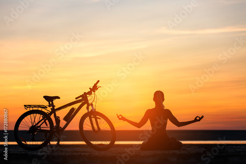 Sports woman doing yoga at sunrise on the sea beach against the background of orange sky and bicycle. Fitness concept. Lotus posture