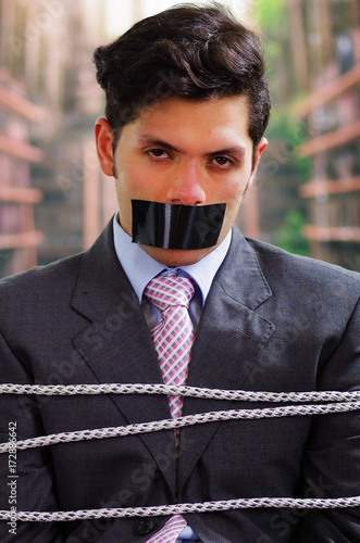 Businessman trapped in a chair with rope, with a black tape in his mouth in a blurred background photo