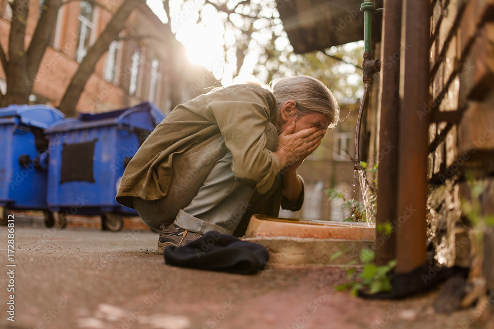 Street life, homeless man squating to wash face. In the yard old tramp with no place of living cleaning face. Street life, homeless man squating to wash face. In the yard old tramp with no place of living cleaning face.