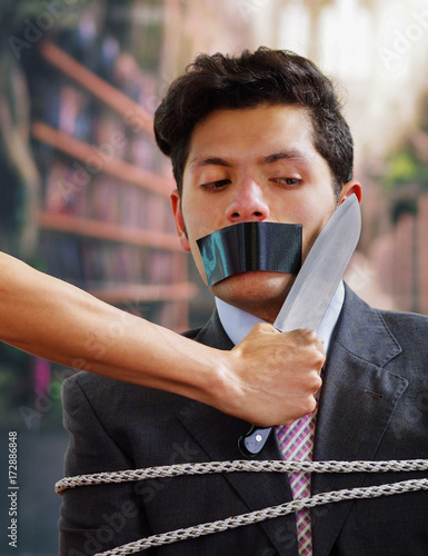 Businessman trapped in a chair with rope, with a black tape in his mouth, afraid of a hand that is holding a knife near of his neack, in a blurred background photo