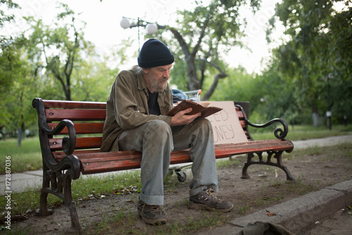 Homeless man sitting on bench holding book in hands. City park, old dirty tramp reading a book.
