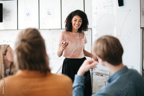 Cheerful African American lady with dark curly hair standing near board and happily looking at her colleagues in office. Young beautiful business woman giving presentation to coworkers during meeting