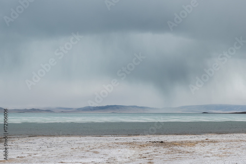 panoramic view of lake with snow on coast and storm clouds in sky 