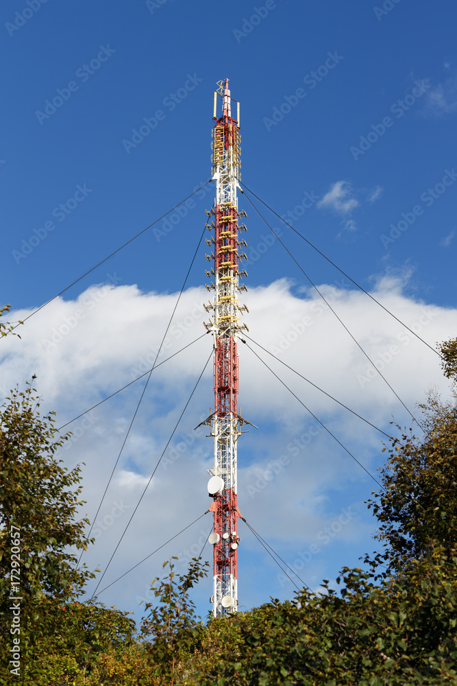 Communications tower with antennas wireless communication channels such a mobile phone tower, cellphone tower, phone pole, surrounded by trees in forest on background blue sky with white clouds.