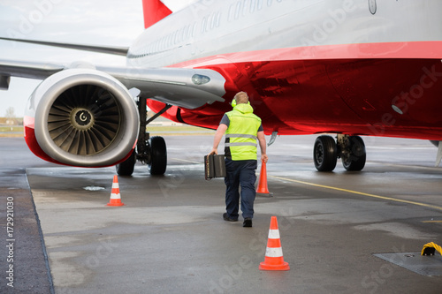 Worker Carrying Traffic Cone And Chocks By Airplane On Runway