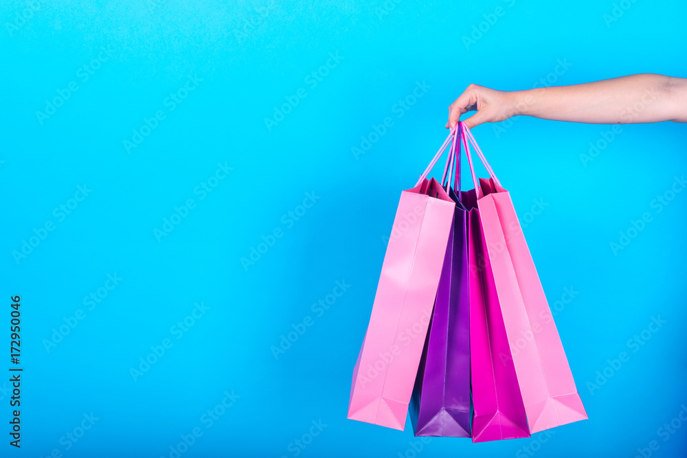 Female hand holding colorful shopping bags isolated on blue (cyan) background, free space, close up. Sale, consumerism, advertisement and black friday concept. Closeup of colored paper shopping bags