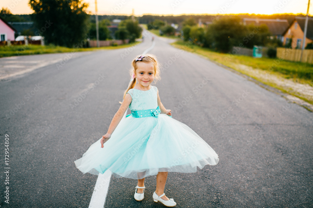 Little beautiful cutie angel girl with lovely face and sincere cheerful eyes posing for camera in aquamarine smart festive holiday fluffy dress with bow. Rural kid on road in countryside at sunset. Little beautiful cutie angel girl with lovely face and sincere cheerful eyes posing for camera in aquamarine smart festive holiday fluffy dress with bow. Rural kid on road in countryside at sunset.