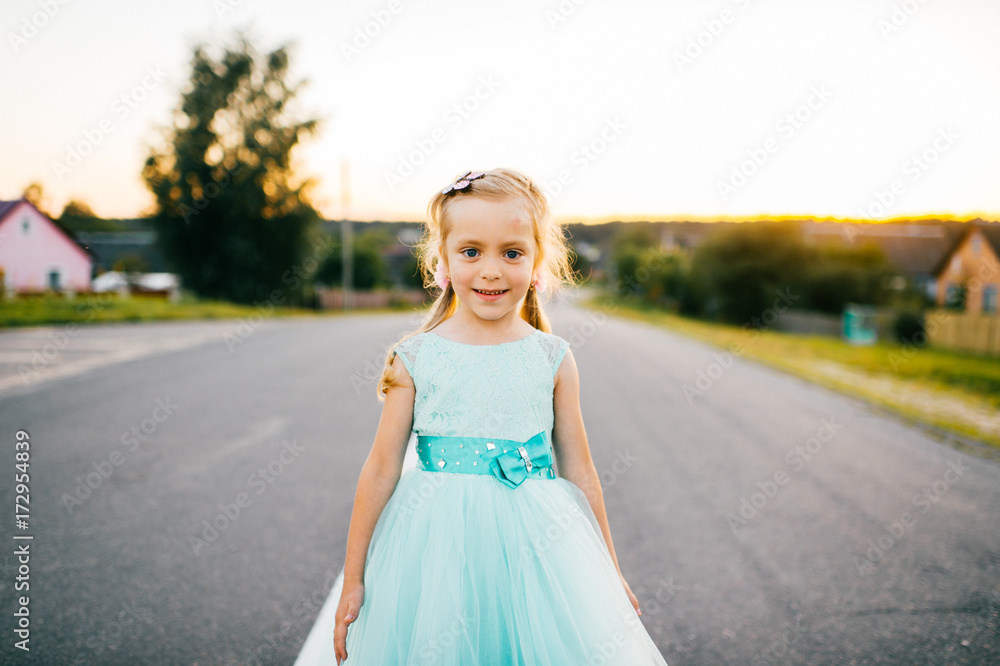 Little beautiful cutie angel girl with lovely face and sincere cheerful eyes posing for camera in aquamarine smart festive holiday fluffy dress with bow. Rural kid on road in countryside at sunset. Little beautiful cutie angel girl with lovely face and sincere cheerful eyes posing for camera in aquamarine smart festive holiday fluffy dress with bow. Rural kid on road in countryside at sunset.