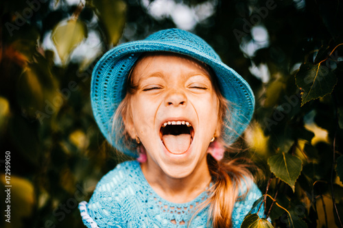 Close up portrait of little beautiful cutie princess girl with emotional face fooling and making faces. Smiling and laughing kid. Funny clown on abstract background in autumn. Orange fall tree leaves