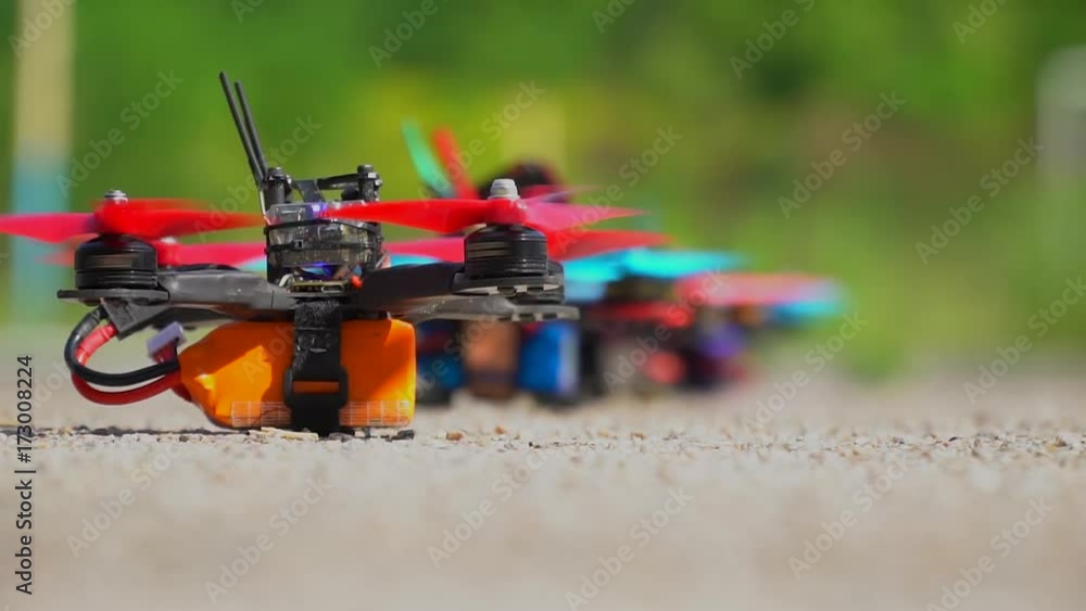 Red and blue racing drone alternately take off from the ground scattering crushed stone in different directions Red and blue racing drone alternately take off from the ground scattering crushed stone in different directions