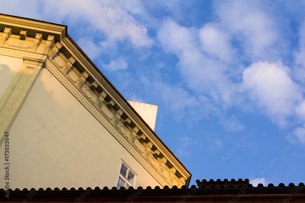 Fototapeta premium Detail of architecture - top of the building with decorated ledge. Blue sky with clouds as copy space. Corrugated pattern of roof tiles at the bottom