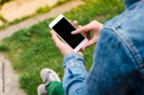 Man use his Mobile Phone outdoor, close up. Mobile phone in hands a young hipster business man in denim shirt and green jeans on the background of green grass.