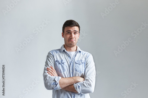 Handsome young male with stubble keeping arms folded, staring at camera with skeptical doubtful facial expression, his look, gesture and grimace expressing disbelief and doubt. Body language