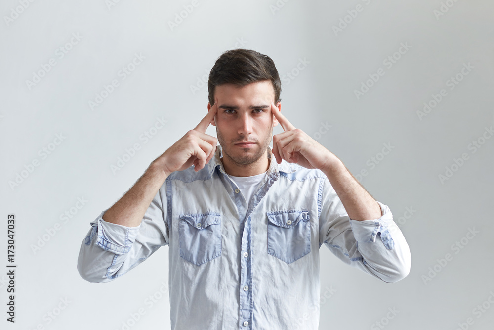 Isolated studio shot of attractive guy in denim shirt squeezing temples with fingers and staring at camera with focused concentrated look. Frustarted male trying hard to remember something important Isolated studio shot of attractive guy in denim shirt squeezing temples with fingers and staring at camera with focused concentrated look. Frustarted male trying hard to remember something important