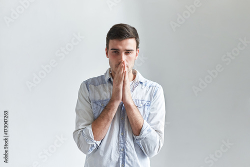 Isolated portrait of thoughtful hopeful young man in denim shirt praying, holding clasped hands at his face and having worried look. Frustrated male facing problem, hoping for the best. Body language