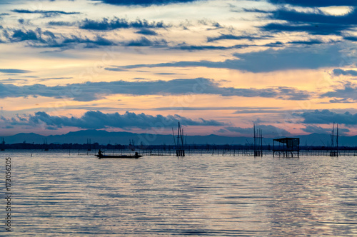Small wooden tropical fishing boat sailing in Songkhla lake during sunset with cloudy twilight sky, fishing cage and fisherman hut.