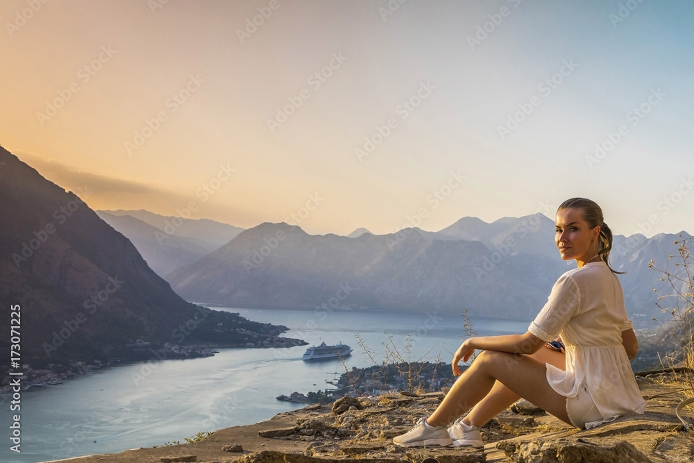 девушка сидит на обрыве скалы с видом на залив и горы в Черногории