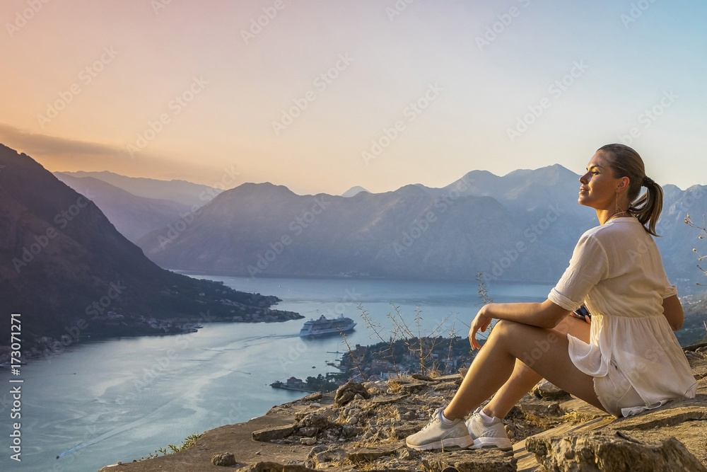девушка сидит на обрыве скалы с видом на залив и горы в Черногории