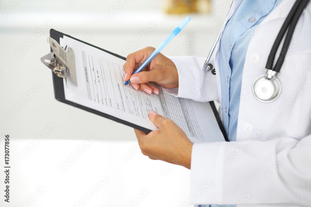 Close-up of a female doctor filling up medical form at clipboard while standing straight in hospital Close-up of a female doctor filling up medical form at clipboard while standing straight in hospital