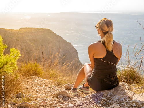 Young women sits at the top of a mountain and looking down on the world. Morning sunrise. Yellow light.