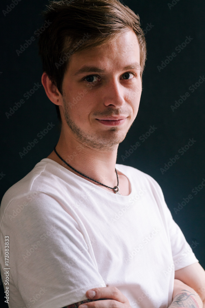Young handsome man in white t-shirt with tattoo on his arm standing near the window looking at the camera. Dark background. Natural daylight. Vertical composition Young handsome man in white t-shirt with tattoo on his arm standing near the window looking at the camera. Dark background. Natural daylight. Vertical composition