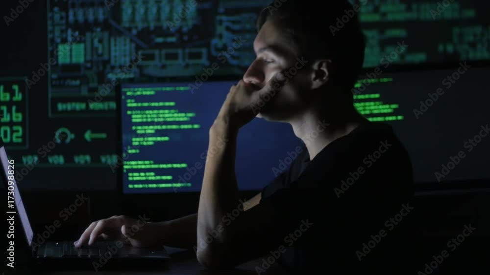 portrait of tired young programmer working at a computer in the data center filled with display screens portrait of tired young programmer working at a computer in the data center filled with display screens