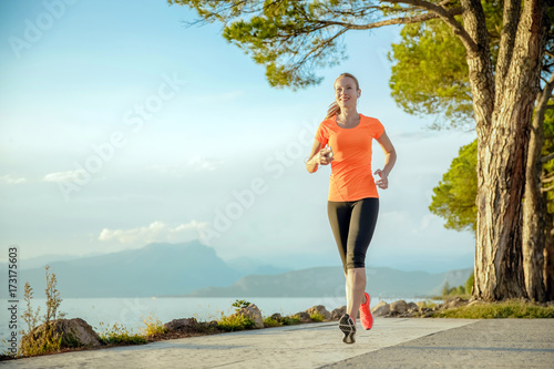Young sexy woman girl is running along the promenade. She is doing her sport workout in the beautiful sundown along the beach. colorful dress, trees, water, mountains and a amazing blue sky.