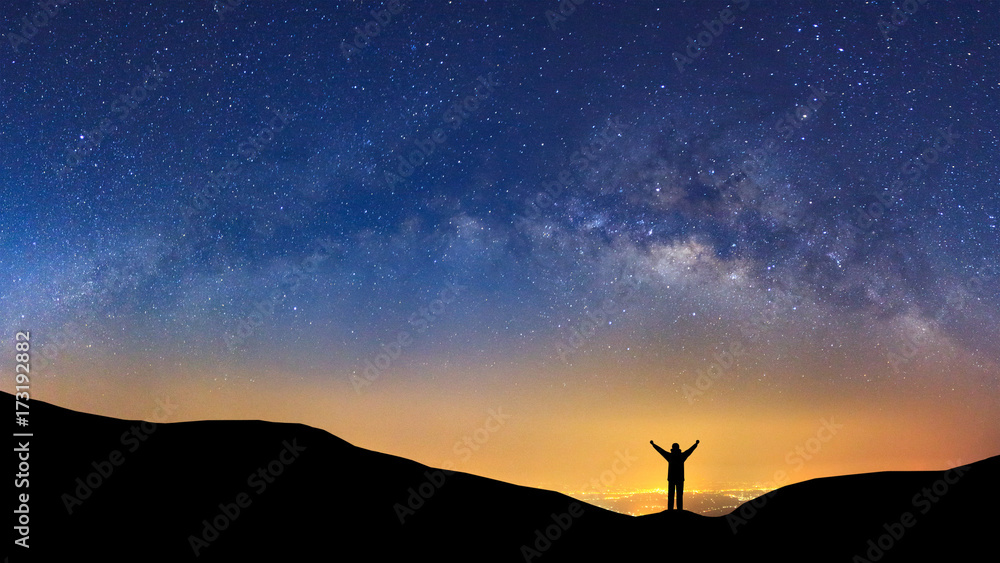 Naklejka premium Panorama landscape with milky way, Night sky with stars and silhouette of a standing sporty man with raised up arms on high mountain.