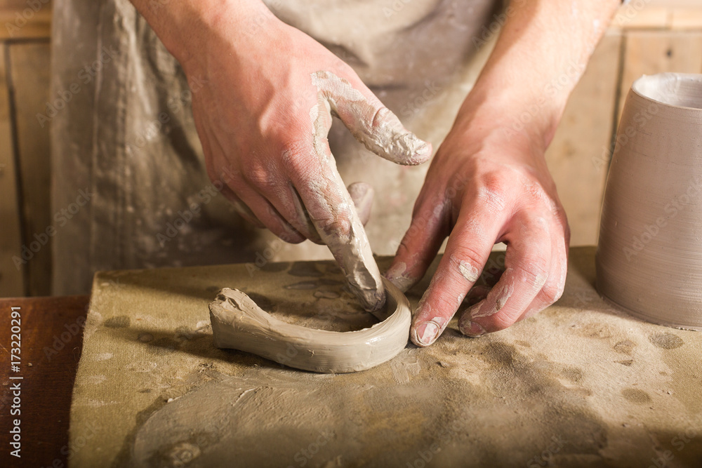 pottery, stoneware, ceramics art concept - closeup on craftsman fingers forming the handle from raw fireclay, master hands work with some piece of clay, male stand at a workshop near the table