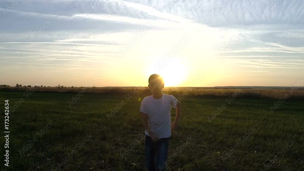 A happy boy in white clothes runs across the green field towards the sun A happy boy in white clothes runs across the green field towards the sun