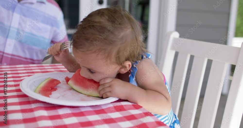 Baby sitting on chair at outdoor table, puts her whole face into a water melon slice to suck on it. Slow motion 4K recorded at 60fps. Baby sitting on chair at outdoor table, puts her whole face into a water melon slice to suck on it. Slow motion 4K recorded at 60fps.