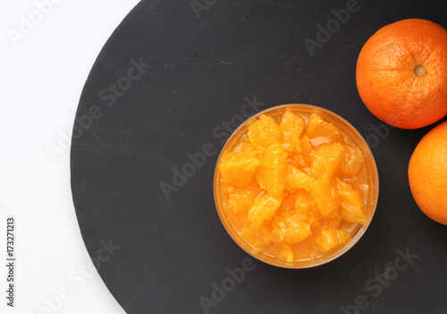 Orange slices in pudding bowl on black round slate with two whole oranges on the side. Top view food background photo