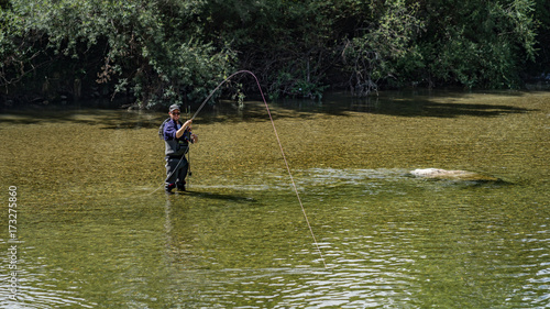 Angler mit Wathose im Wasser beim Angeln mit Fliegenrute bei Sonne im klaren Fluss stehend und werfend