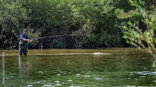 Angler mit Wathose und Fliegenrute im Wasser beim Angeln bei Sonne im klaren Fluss stehend und werfend