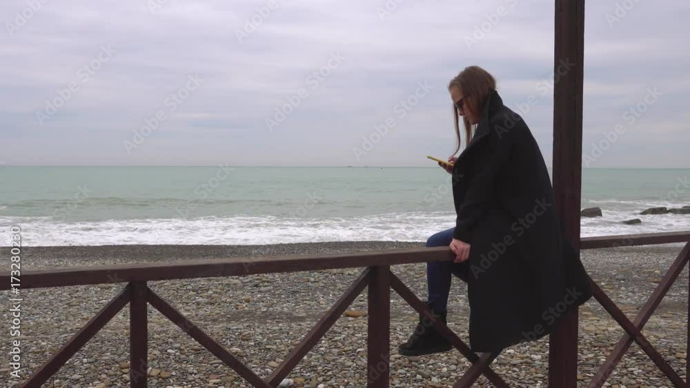 Caucasian woman in black coat sits on wooden railing on a cold sea beach. Handheld shot Caucasian woman in black coat sits on wooden railing on a cold sea beach. Handheld shot